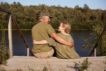 Fototapeta premium Father and son fishing together, sitting on wooden placement on bank of river, hugging and looking at each other, wearing casual attires, spending sunny day in open air.