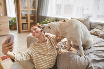 Obraz na płótnie Canvas Warm toned portrait of happy young woman taking selfie with dog giving kisses in cozy home interior lit by sunlight, copy space