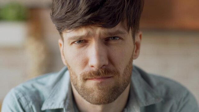 Head and shoulders closeup portrait of sad young man with beard raising eyes and looking at camera with unhappy frowning facial expression