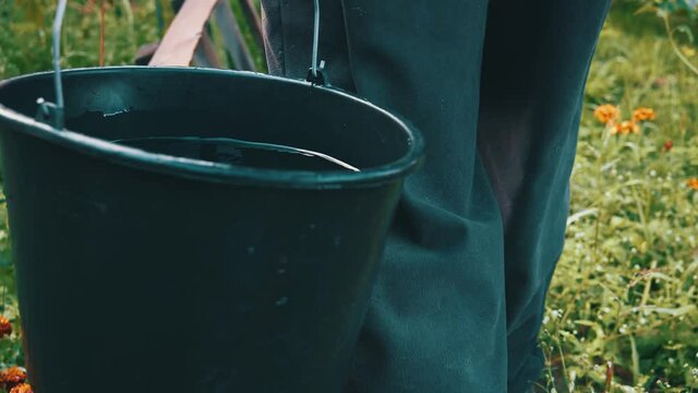 Man keeping plastic bucket with water while walking at the path around bushes and plants. Unrecognizable man walking at the village. Nature and summer concept