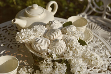 Soft and sweet marshmallows on the beautiful plate. White lilac flowers on the vintage table. Teapot and cups are ready for breakfast at the garden. Bright spring mood.