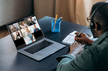 Fototapeta na wymiar Distance education, online lesson. African American guy in headset learns remotely and takes notes using laptop and video chatting app. On the laptop screen the teacher and group of diverse people