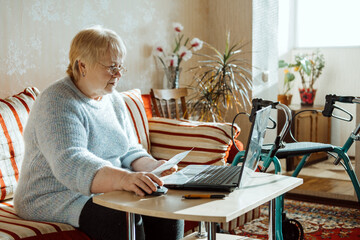 Fototapeta na wymiar Life Insurance, Disability medical insurance policy for Seniors. Mature woman in glasses with Laptop and documents on the background of Rollator Mobility Walker for Seniors at home.