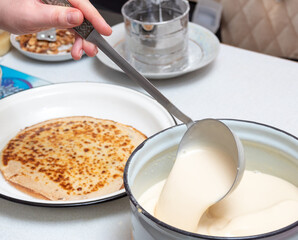 A hand holds a metal ladle while stirring the pancake batter. In the background is already baked pancake. Preparation for the celebration of Maslenitsa. Selective focus.