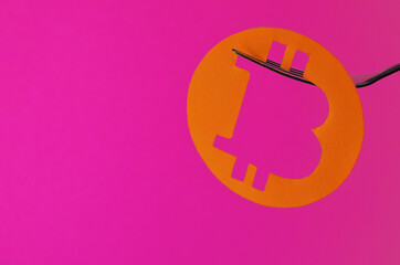 A bitcoin coin carved from orange paper hangs on a fork on a pink background with a copy space. Conceptual collage about cryptocurrency in the style of minimalism. High quality photo