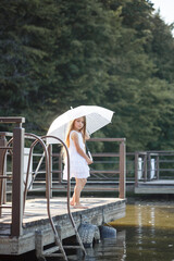 A beautiful girl of six years old in a short white dress stands on the masonry near the river and holds a white umbrella in her hands
