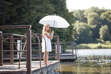Fototapeta na wymiar A beautiful girl of six years old in a short white dress stands on the masonry near the river and holds a white umbrella in her hands