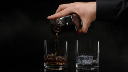 Hand of barman pouring golden whiskey, cognac or brandy from bottle into glasses with ice cubes on...