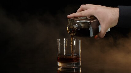 Barman pouring golden whiskey, cognac or brandy from bottle into glass with ice cubes on black background. Bartender pouring glass of alcohol drink bourbon, rum. Shoot on cinema camera. Copy-space