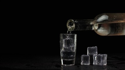Bartender pouring up shot of vodka with ice cubes from bottle into drinking glass against black background. Barman pour clear transparent alcohol drink vodka tequila in shot-glass. Copy-space