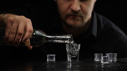 Alcoholic guy drinking cold transparent alcohol drink sake, tequila or rum. Man pouring up frozen vodka from bottle into shot glass with ice cubes on black background. Alcohol dependence addiction