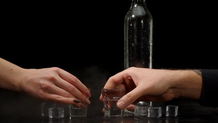 Bartender pouring up two shots of vodka with ice cubes from a bottle into glasses against black background. Two hands with glasses of vodka making cheers. Raising toast with glasses of tequila or sake
