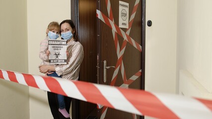 Sick mother with kid daughter in medical masks holding inscription text, opening entrance corridor door with warning sign. Family of woman and child girl during coronavirus quarantine lockdown at home