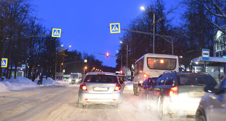 Winter frosty evening. Cars are driving along the snow-covered road. On both sides of the road there are bus pavilions and traffic signs: a pedestrian crossing and a bus stop.
