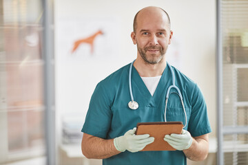 Waist up portrait of mature male veterinarian smiling at camera while holding cliboard at vet...