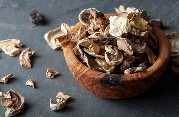 Close up isolated angled image of a rustic wooden bowl filled with sliced and dried mushroom pieces. The bowl is on dark stone surface. There is an assortment of mushroom types in this moody image.