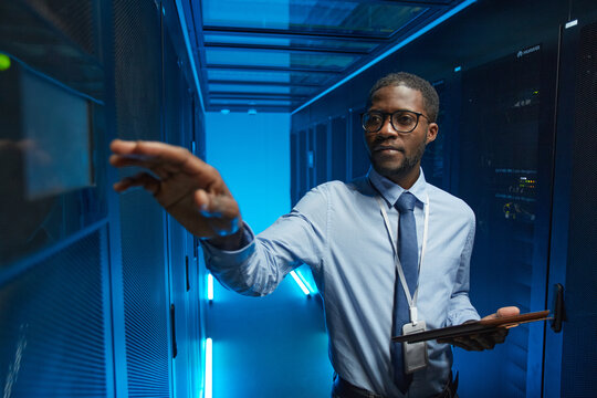 Waist up portrait of African American man reaching for server cabinet while working with supercomputer in data center and holding tablet, copy space