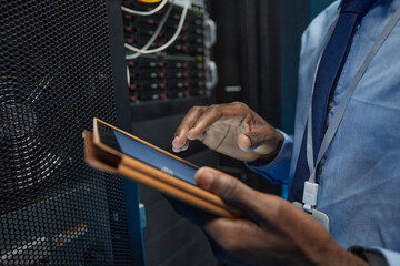 Close up of African American man holding digital tablet while standing by server cabinet and working with supercomputer in data center, copy space