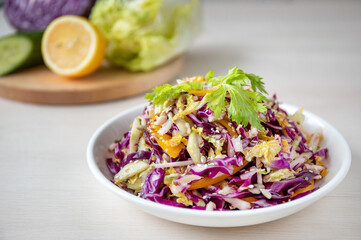 Cucumber, red cabbage, lemon on the board and vegetable salad in a white plate are on the table.