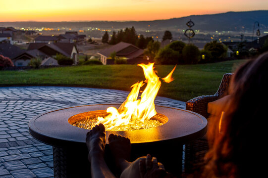 A woman relaxes by a roaring firepit on a paver patio at sunset overlooking the Spokane Valley, in Washington State, USA