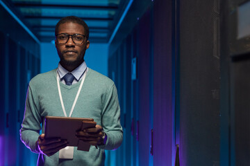 Fototapeta na wymiar Waist up portrait of African American data engineer looking at camera while working with supercomputer in server room lit by blue light, copy space
