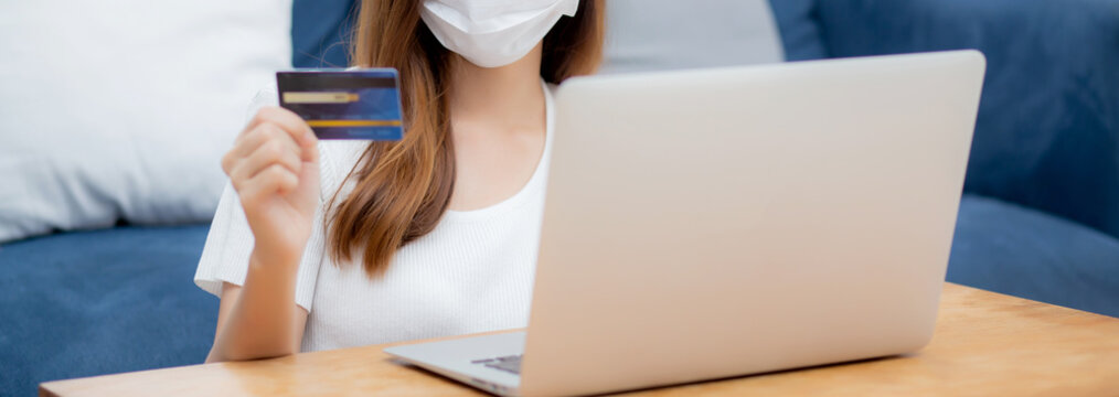 Young Asian Woman In Face Mask Holding Credit Card Shopping Online With Laptop Computer Buy And Payment, Girl Using Debit Card Purchase Or Transaction Of Finance, Lifestyle And New Normal Concept.