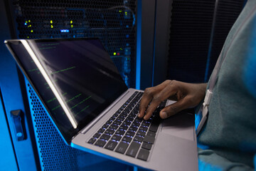 Close up of young African American data engineer holding laptop while working with supercomputer in server room lit by blue light, copy space