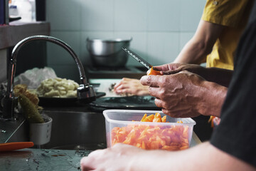 Las manos de un hombre con un cuchillo y una zanahoria siendo cortada en partes pequeñas dentro de un recipiente de plástico en la cocina mientras otra persona de fondo prepara un pulpo
