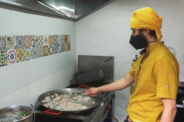 Un hombre con tapabocas y pañoleta amarilla cocinando camarones en una olla de un fogón