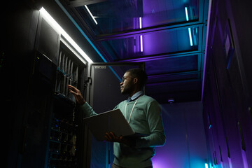 Low angle portrait of young African American data engineer working with supercomputer in server room lit by blue light and holding laptop, copy space - obrazy, fototapety, plakaty