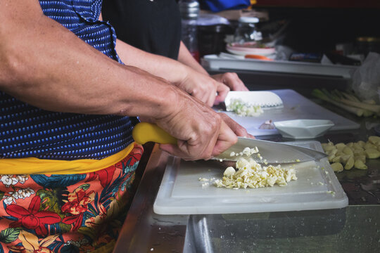 Las manos de un hombre picando en una tabla con un cuchillo y de fondo elementos de cocina y oficina y una mujer cortando cebolla con otro cuchillo