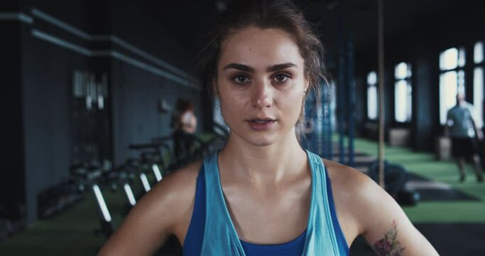 Portrait of serious beautiful young determined female athlete looking at camera, sweaty after exercising at local gym.