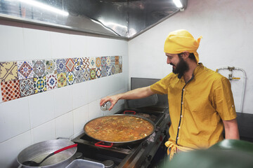 Un hombre delgado de barba y ojos pequeños con pañoleta amarilla sonríe mientras echa condimentos a una paella en una olla dentro de la cocina