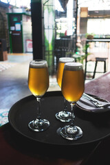 Tres copas de vidrio con cerveza servidas en una bandeja tipo charol con un tenedor y una servilleta al lado y de fondo una silla, un mirador, un local y una reja