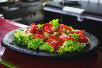 Una ensalada de lechuga, tomate y aceitunas mientras cae una salsa sobre ella, servida en un plato de barro y de fondo se ven un fogón y un refrigerador