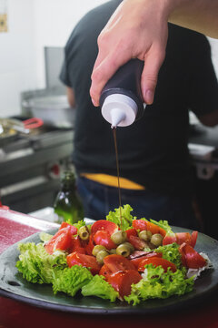 Una ensalada de lechuga, tomate y aceitunas mientras la mano de un hombre esparce una salsa sobre ella, servida en un plato de barro y de fondo se ven un fog&oacute;n y otro hombre