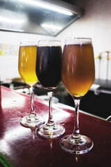 Tres copas de vidrio con cervezas de diferentes colores y sabores en una barra, de fondo se ven un fogón y un refrigerador