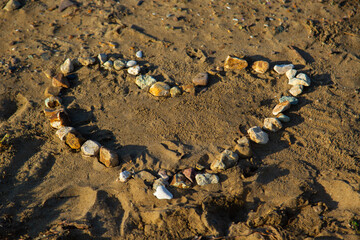 Fototapeta na wymiar Heart laid out of sandy multi-colored stones on yellow sand with lateral sun. Minimalism at sea.
