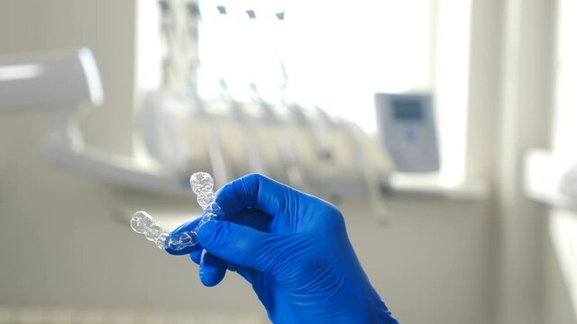Modern transparent aligners or braces to straighten teeth in cosmetic dentistry and orthodontics. Female hand in blue glove holding plastic aligner for bite correction. Dental equipment background. 4