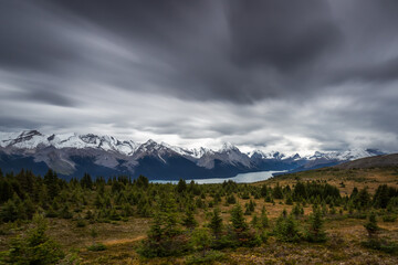 Long exposure of a Canadian Rockies landscape with a lake next to mountains with snow during autumn under a looming overcast sky with fast dark clouds, Maligne Lake, Jasper National Park, Canada