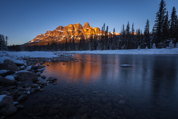 Calm Canadian Rockies winter landscape at sunset of a cold river with rocks, snow, ice, trees and orange mountains reflected in the clear water, Castle Mountain, Banff National Park, Alberta, Canada