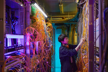 Wide angle side view at young network technician connecting wires and cables in server room, copy...