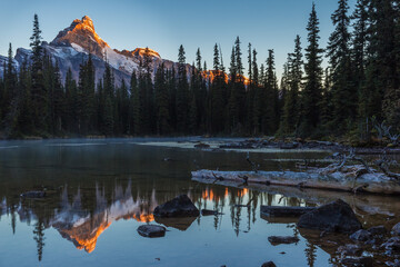 Serene autumn sunrise, warm orange light glowing on mountain peak with snow in a cold morning, reflections on still water of a small pond with rocks and trees, Yoho National Park, BC, Canada 