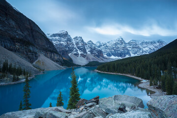 Sunrise in the mountains with snow during a cloudy fall morning, blue calm lake with reflections, green trees and gray rocks, Valley of the Ten Peaks, Moraine Lake, Banff, Alberta, Canada
