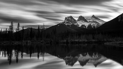 Black and white long exposure of three mountains (Three Sisters) with snow, reflected on a pond under a grey sky with fast clouds during fall, Canmore, Alberta, Canada