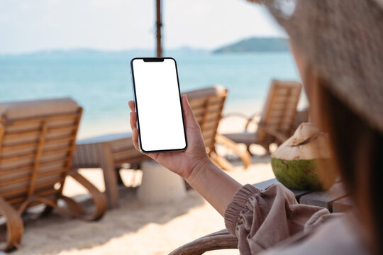 Mockup image of a woman holding mobile phone with blank desktop screen while sitting on the beach