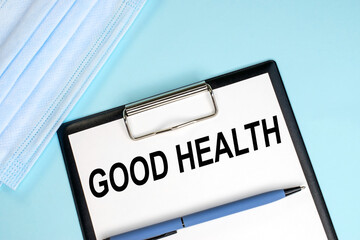 Good health is written on a stationery tablet that lies next to a medical face mask on a blue background. Medical concept