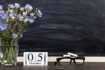 Flowers in a glass cup, glasses and a calendar with the date of October 5, against the background of chalkboard, Teacher's Day concept. Copy space.