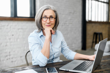 Intelligent mature senior female business owner in glasses with hand on a chin looking at the...