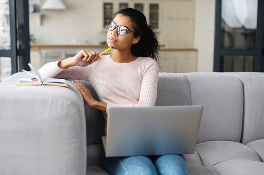 Thoughtful Multiracial Young Woman With Dark Ponytail, Sitting On The Sofa With Laptop And Looking Away, Holding Pencil In The Hand Near The Face, Thinking About Plans, Ideas, Planning A Purchase